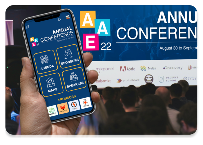 A hand holding a mobile event app featuring an event homescreen in front of an audience watching a conference presentation on stage.