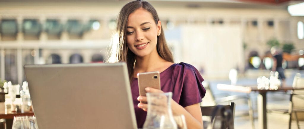 Smiling woman in a terrace sitting at a dinner table, with cups and a laptop,  holding a mobile phone.